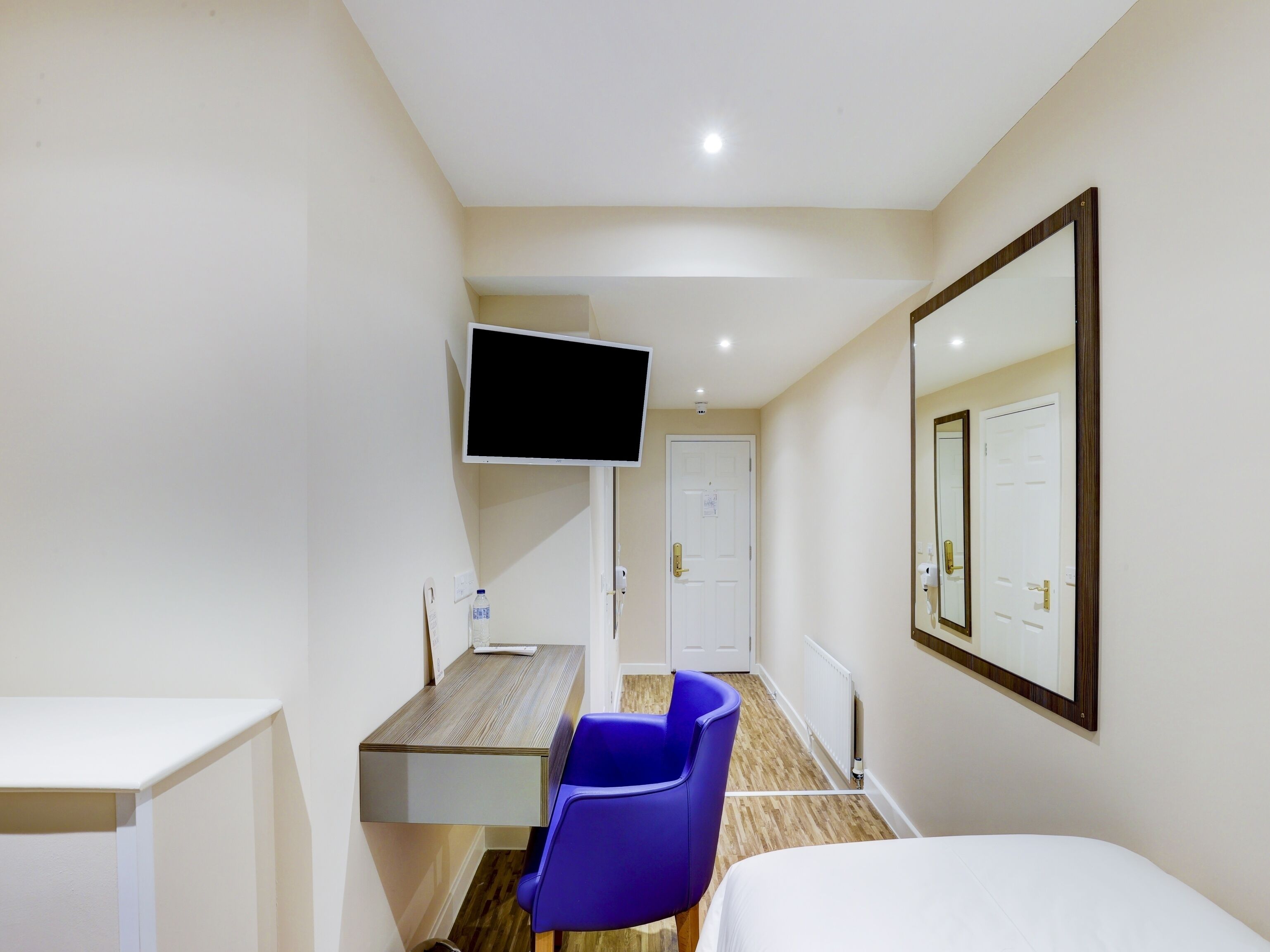 A hotel room with a bed, a desk with a chair, a television mounted on the wall, and a door.