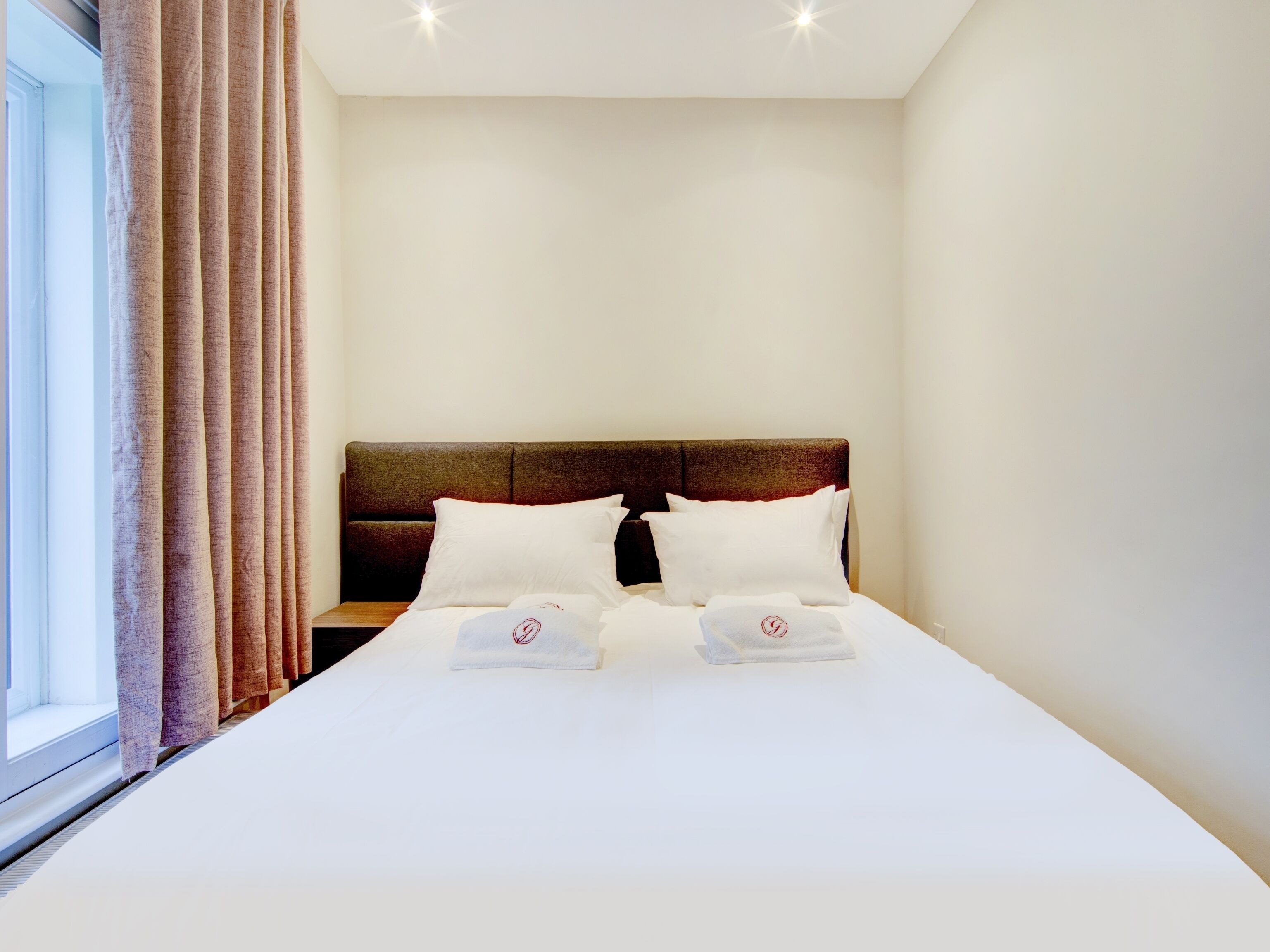 Un lit bien fait, avec des draps blancs et deux oreillers, une tête de lit en bois et une fenêtre avec des rideaux.