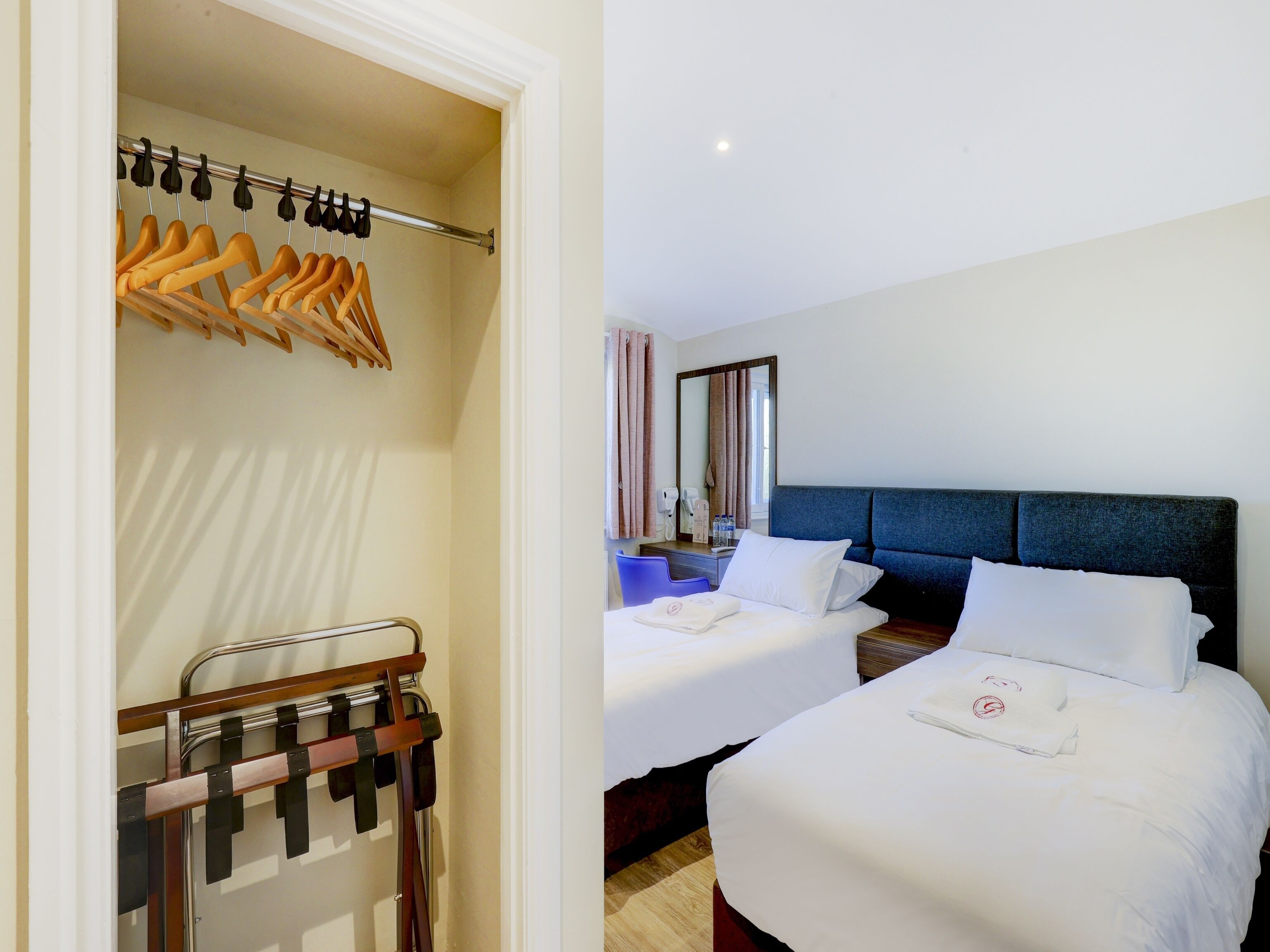 A hotel room with two beds, a wardrobe with hanging clothes, a bathroom with a mirror, and a bedside table with toiletries.