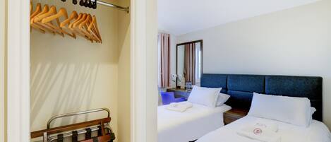 A hotel room with two beds, a wardrobe with hanging clothes, a bathroom with a mirror, and a bedside table with toiletries.