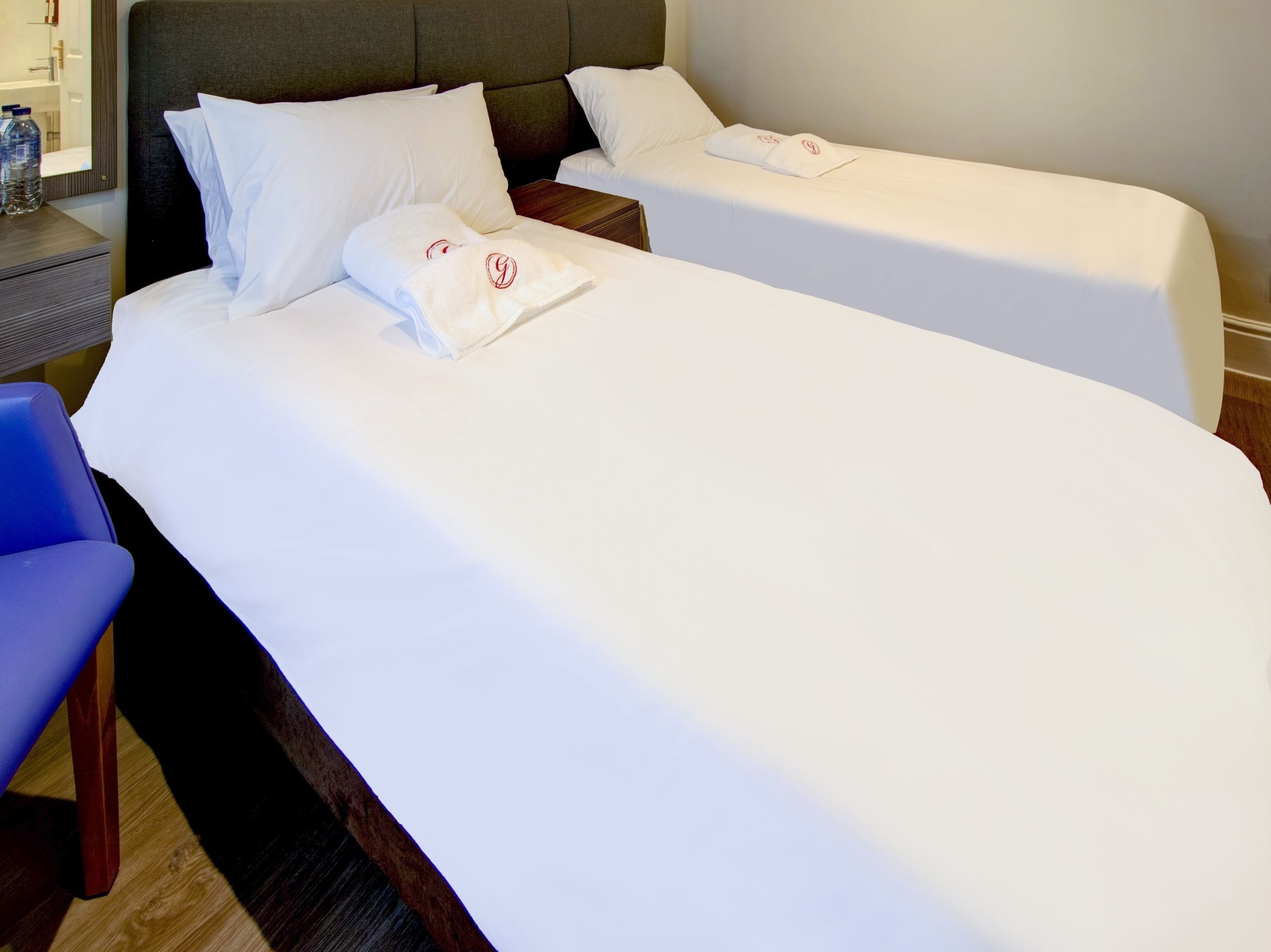 A hotel room with two beds, each with white linens and pillows, a wooden nightstand, a blue chair, and a small table with a bottle of water.