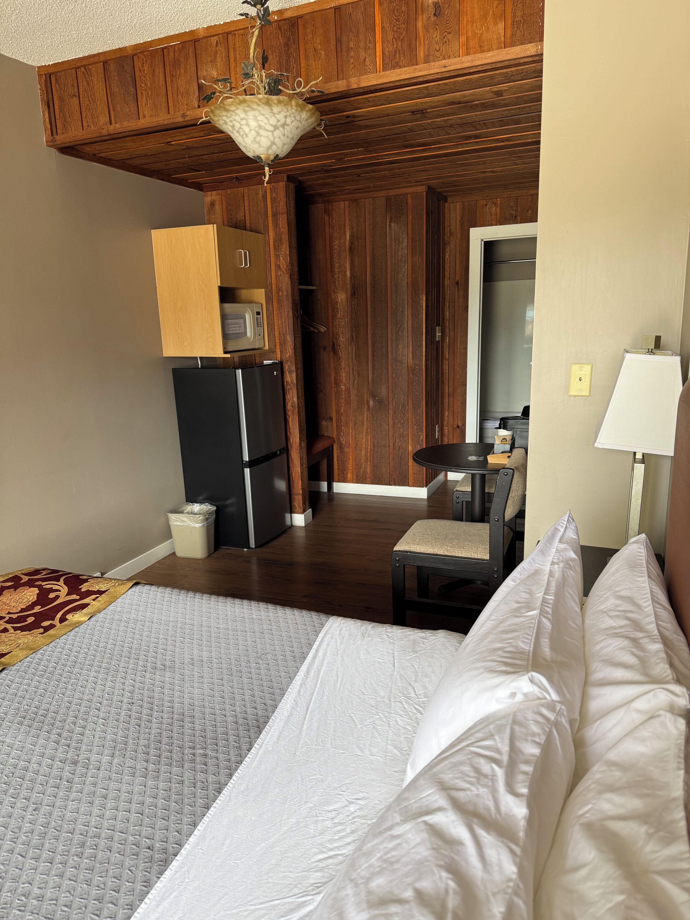 A hotel room with a bed, a refrigerator, a microwave, a wooden ceiling, and a small table with a chair.