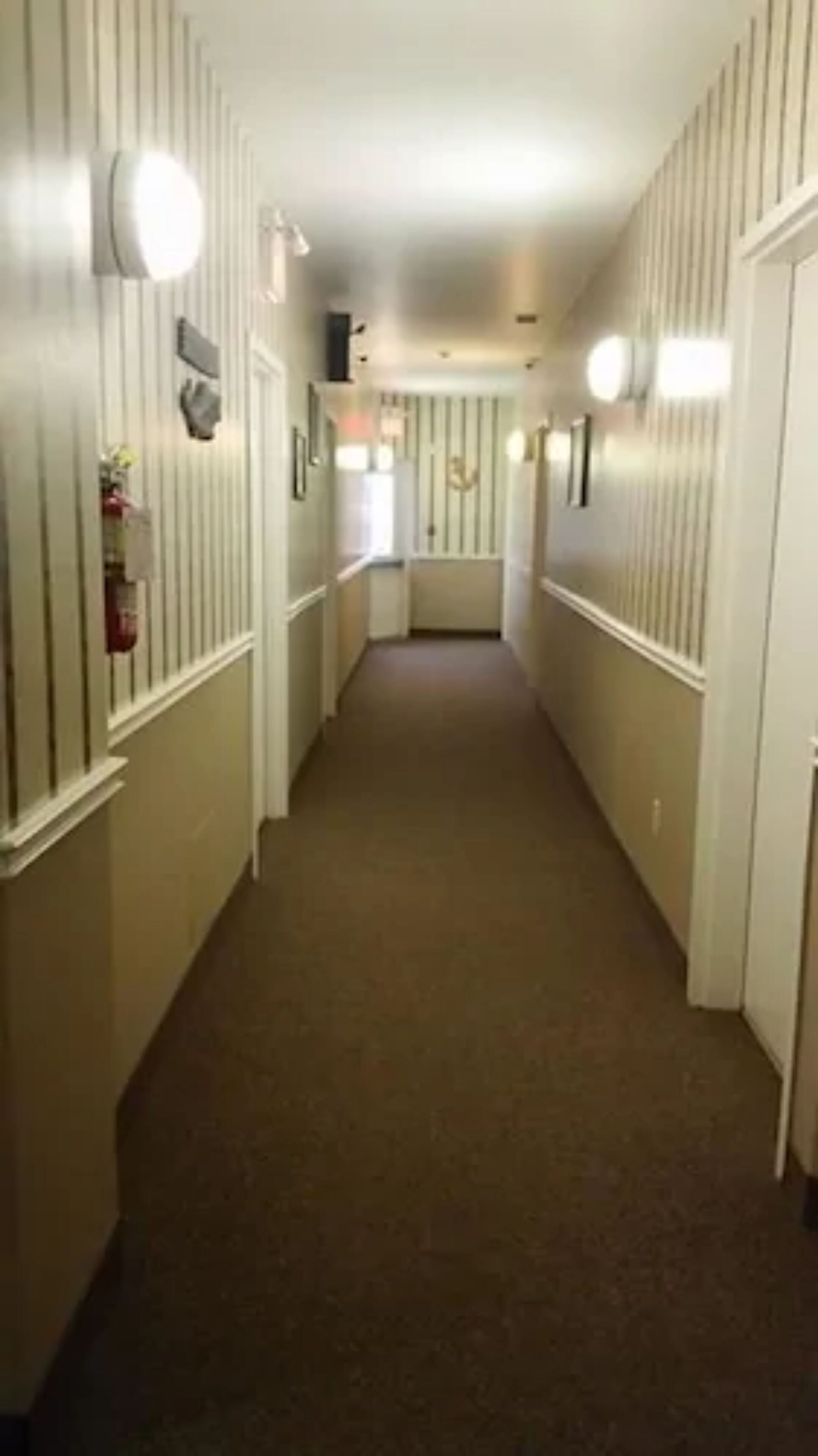 A narrow, carpeted hallway with white walls and vertical stripes, leading to a door at the end.
