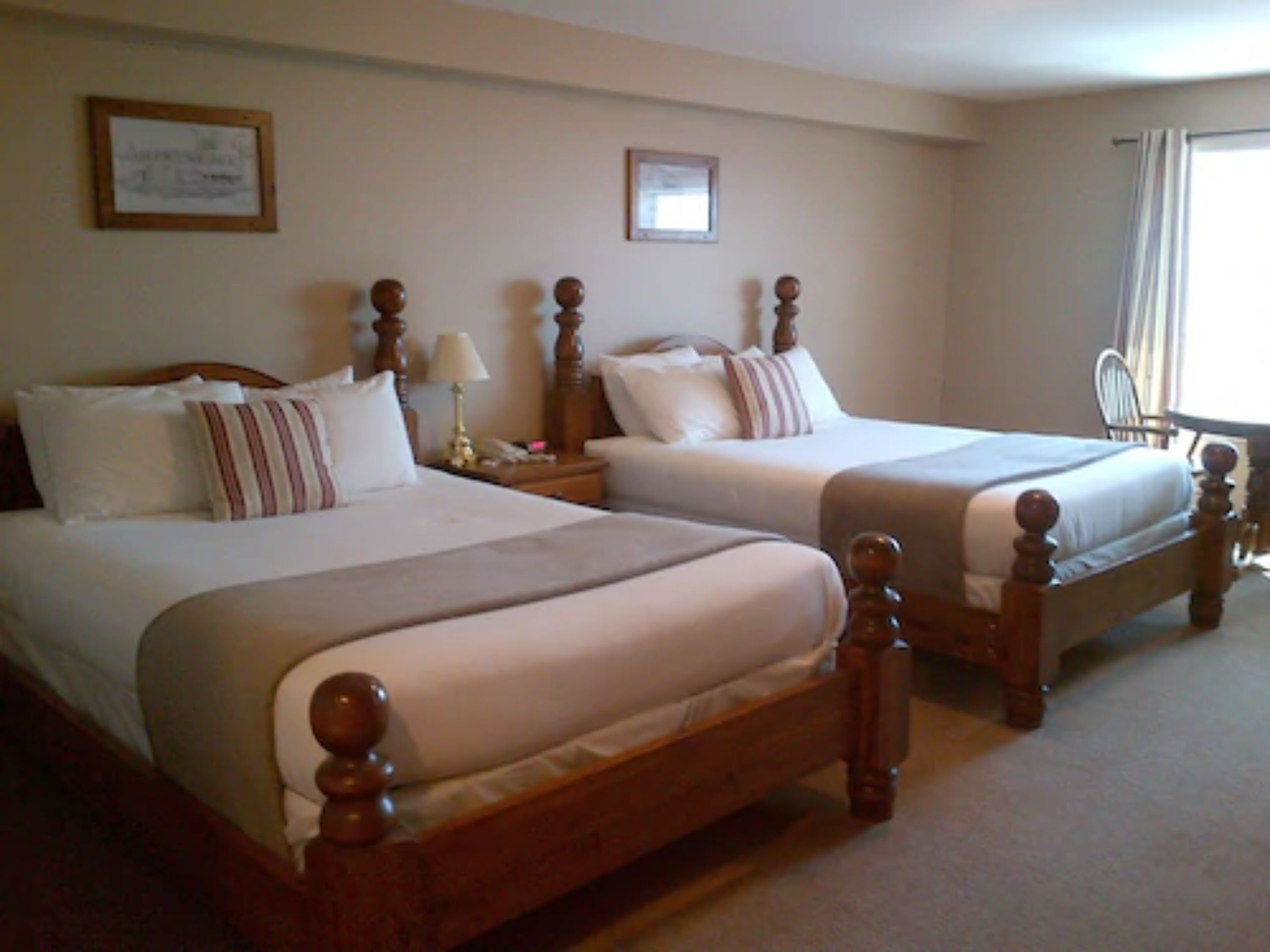 Habitación de hotel con dos camas dobles, cabeceros de madera, una mesita de noche con lámpara y una silla cerca de la ventana.