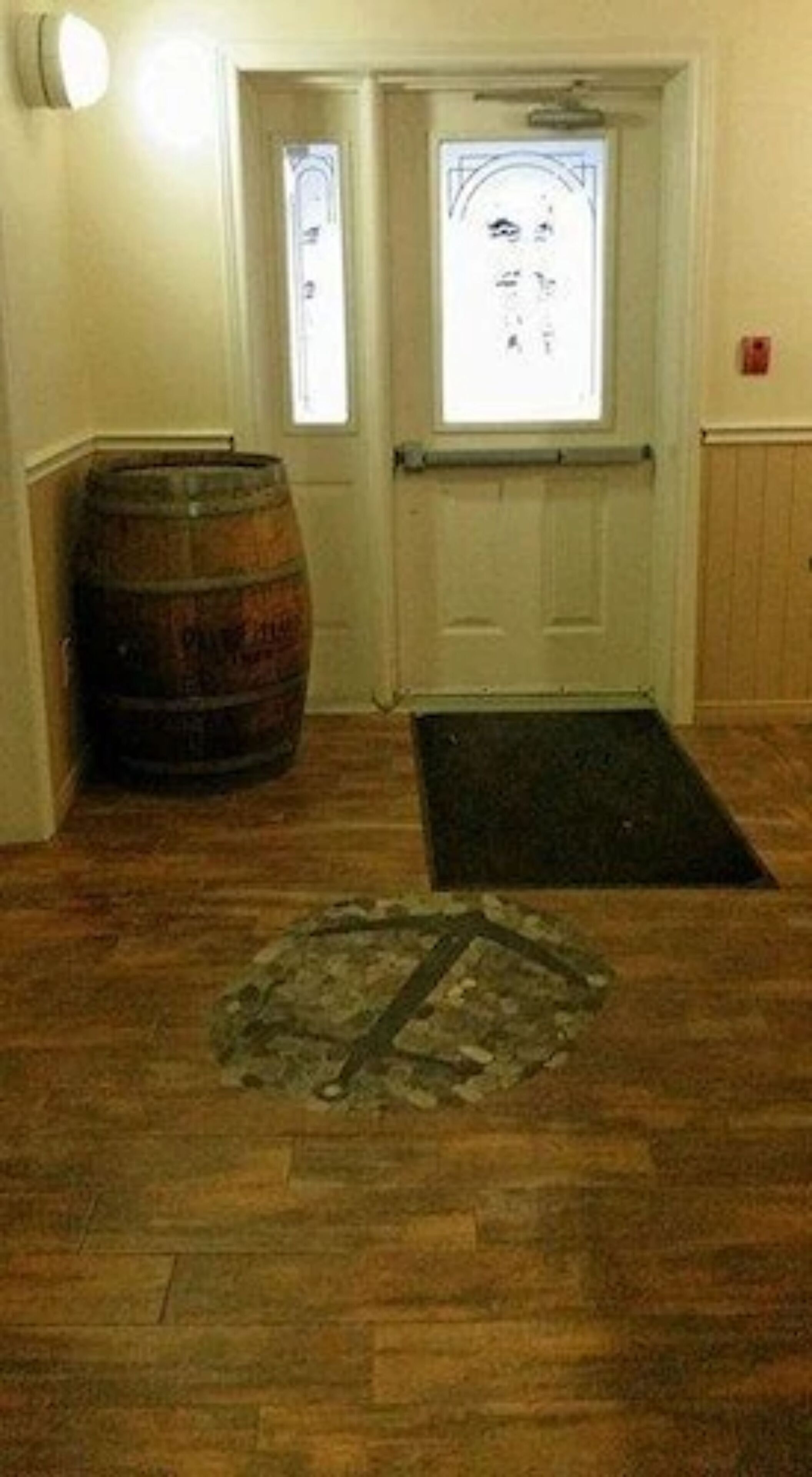 Une pièce avec un plancher en bois, une barrique en bois, une porte avec une fenêtre et un paillasson à l’entrée.