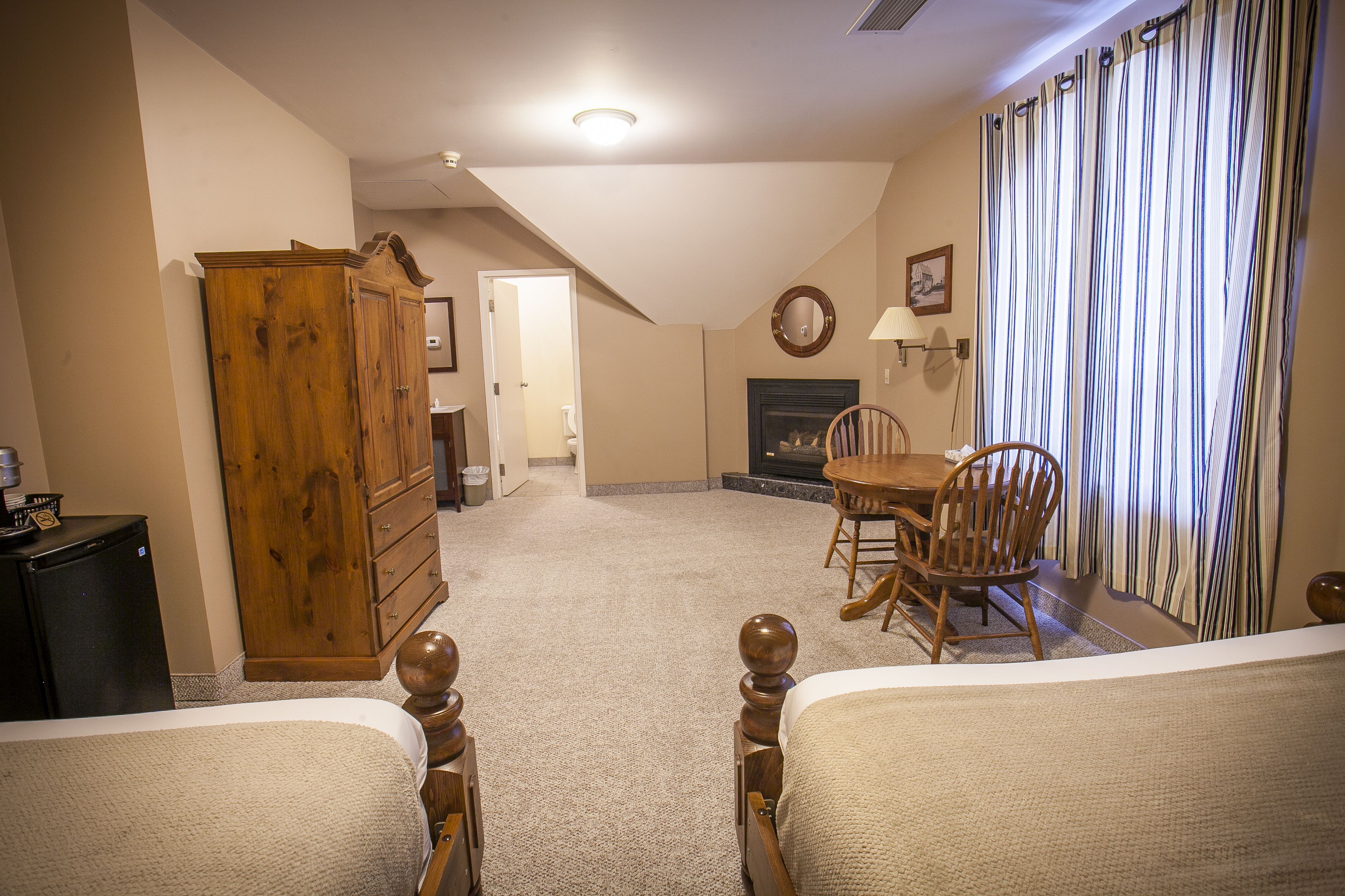 A bedroom with a large wooden wardrobe, a fireplace, a dining table with chairs, and a view of a striped curtain.