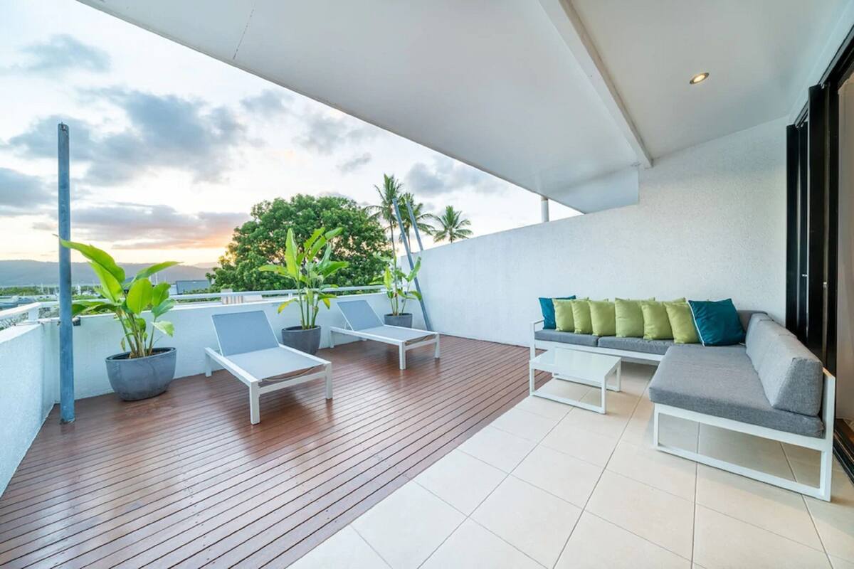 Ein Balkon mit Holzboden, weißen Lounge-Stühlen, einem grauen Sofa mit grünen und blauen Kissen und Topfpflanzen.