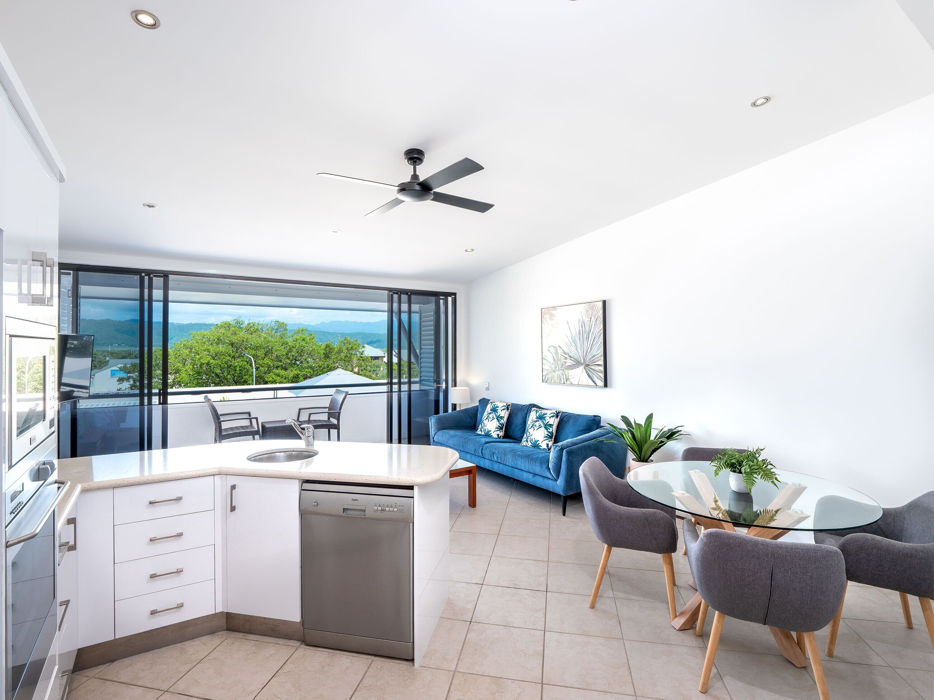 A modern kitchen with a dishwasher, a dining area with a glass table and chairs, a blue sofa, and a view of the outdoors.