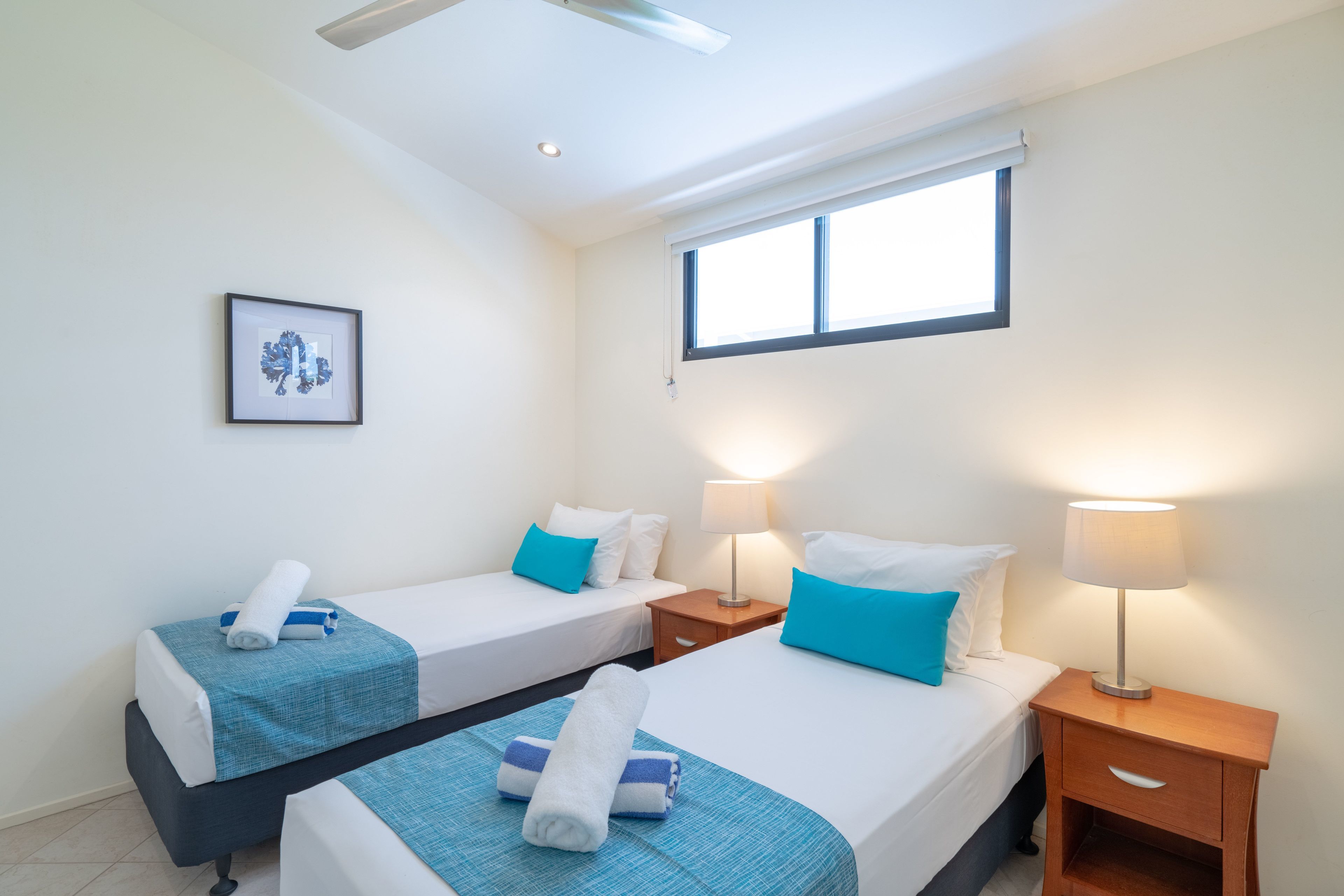 Une chambre d’hôtel avec deux lits, chacun recouvert d’une couvre-lit bleue et doté de draps blancs, une table de chevet avec une lampe, une œuvre d’art encadrée au mur et une fenêtre avec des stores.