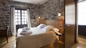Un dormitorio con pared de piedra, puerta de madera, cama con ropa blanca y una silla.