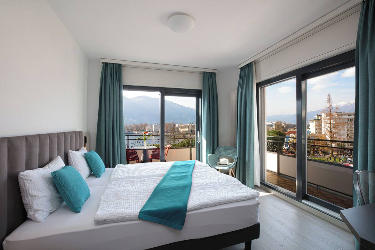 Ein Hotelzimmer mit einem großen Bett, Blick auf die Stadt und Berge sowie einem Balkon mit Tisch und Stühlen.