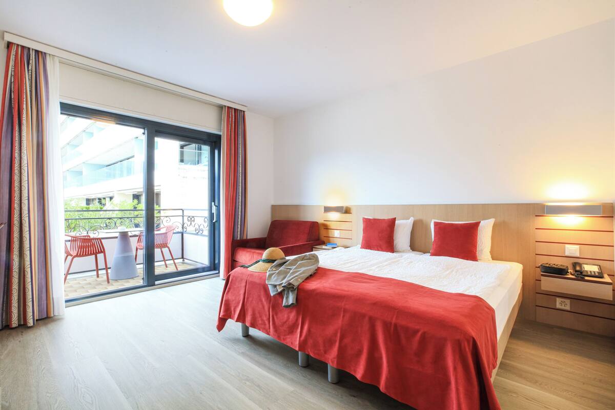 Ein Hotelzimmer mit einem großen Bett, einem Balkon mit roten Stühlen und Blick auf Gebäude.