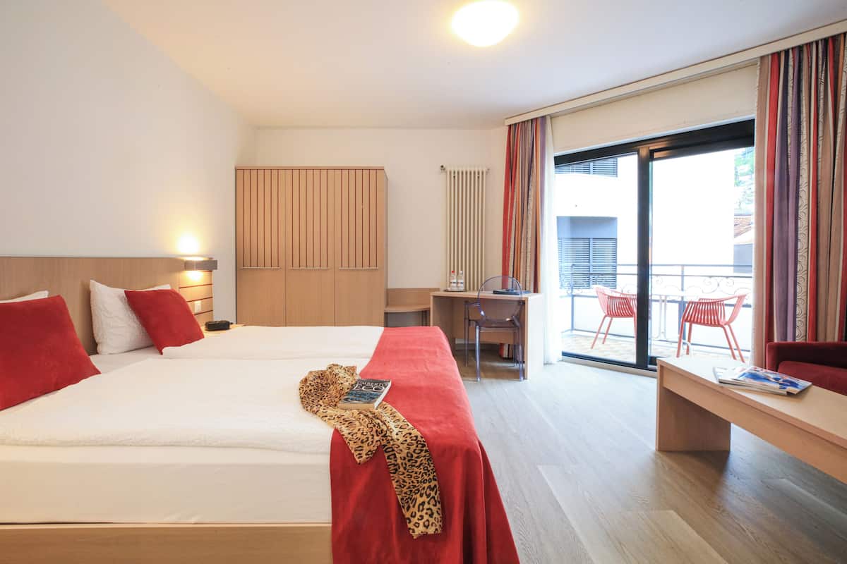 Ein Hotelzimmer mit zwei Betten, einem Schreibtisch und einem Balkon mit roten Stühlen.
