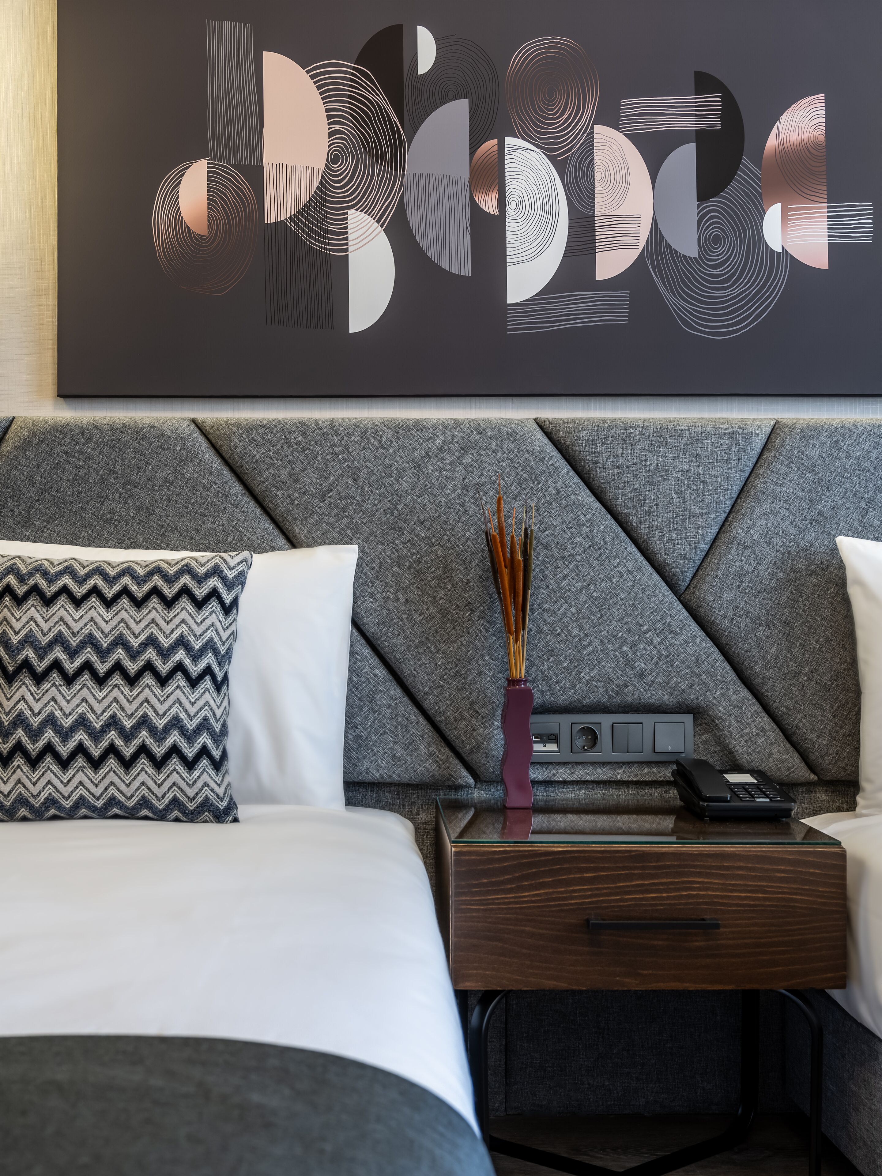 Una habitació d'hotel moderna amb una gran obra d'art abstracta a la paret, un llit ben arrenjat amb almoinades estampades, una taula de nit amb un ram i un telèfon, i una estació de càrrega penjada a la paret.