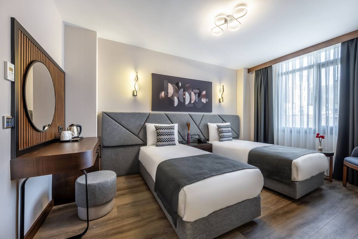 A modern hotel room with two beds, a large mirror, wall art, and a view of the outdoors.