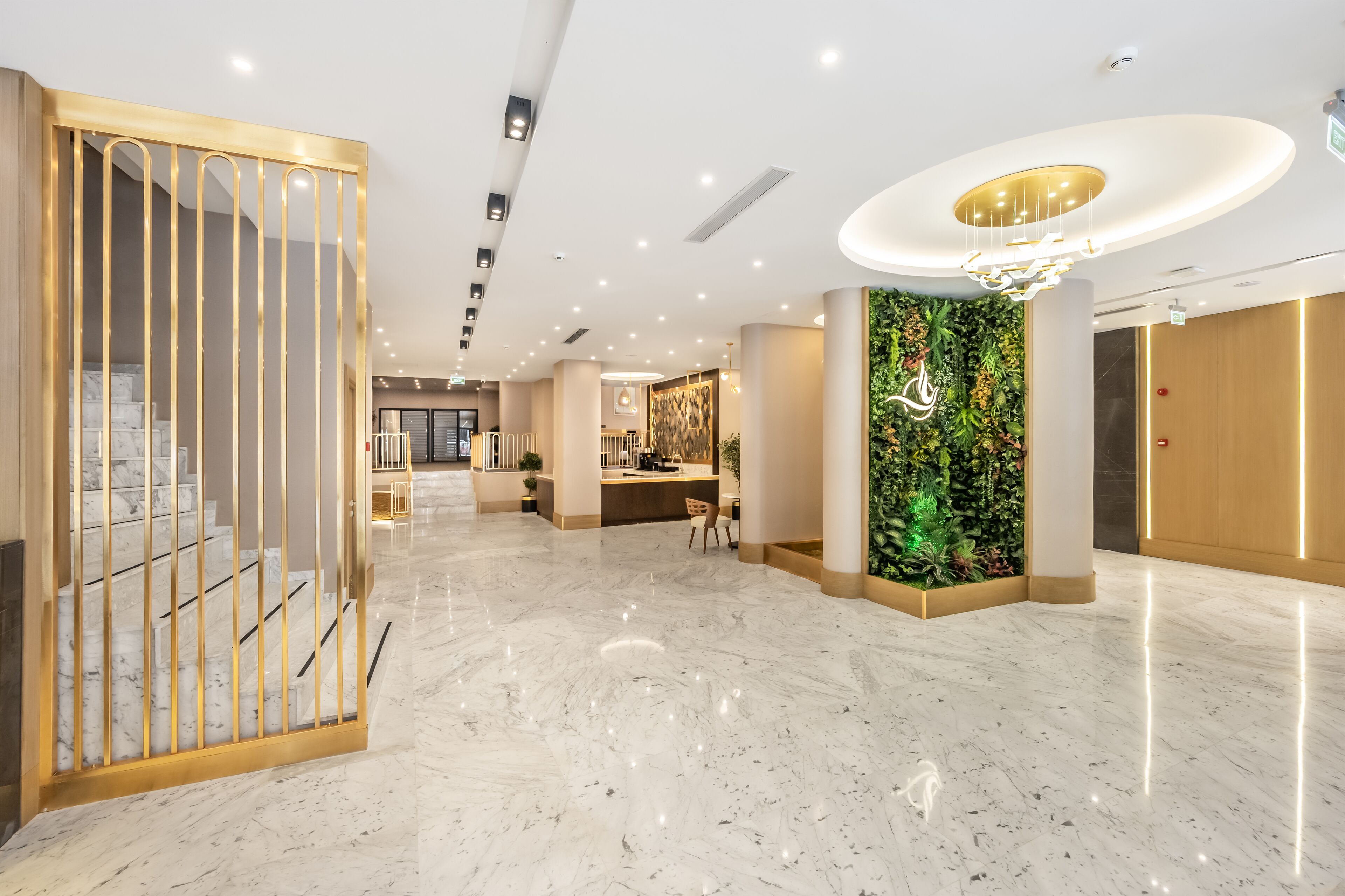 Eine moderne Hotelhalle mit Marmorboden, einer Rezeption, einer Treppe mit goldfarbenen Geländern und einer vertikalen grünen Wand.
