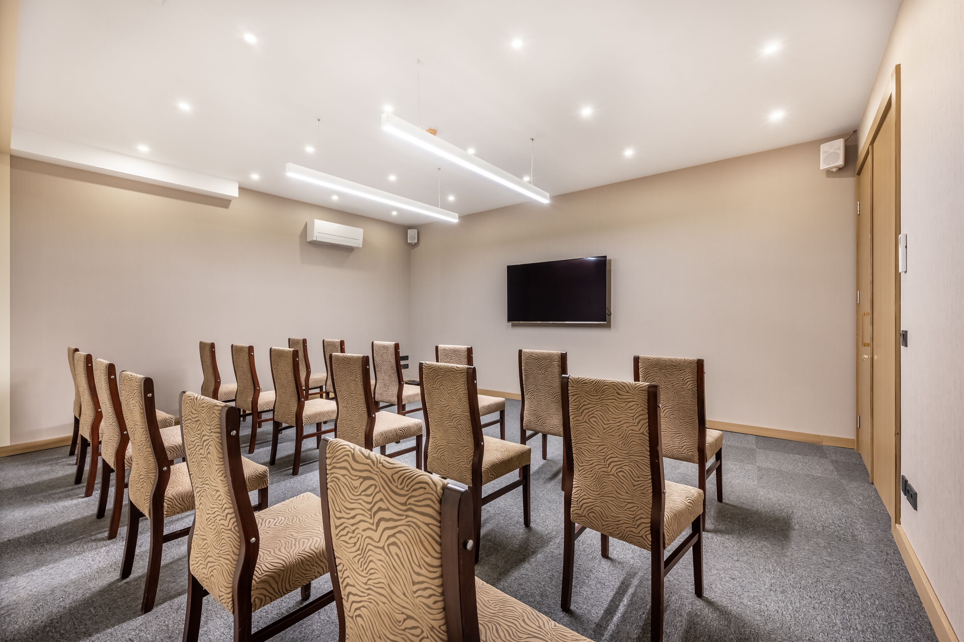 A conference room with rows of chairs, a flat-screen TV on the wall, and recessed ceiling lights.