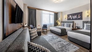 Une chambre d’hôtel moderne équipée d’un canapé, de deux lits, d’une télévision, d’un bureau et d’une grande fenêtre avec des rideaux.