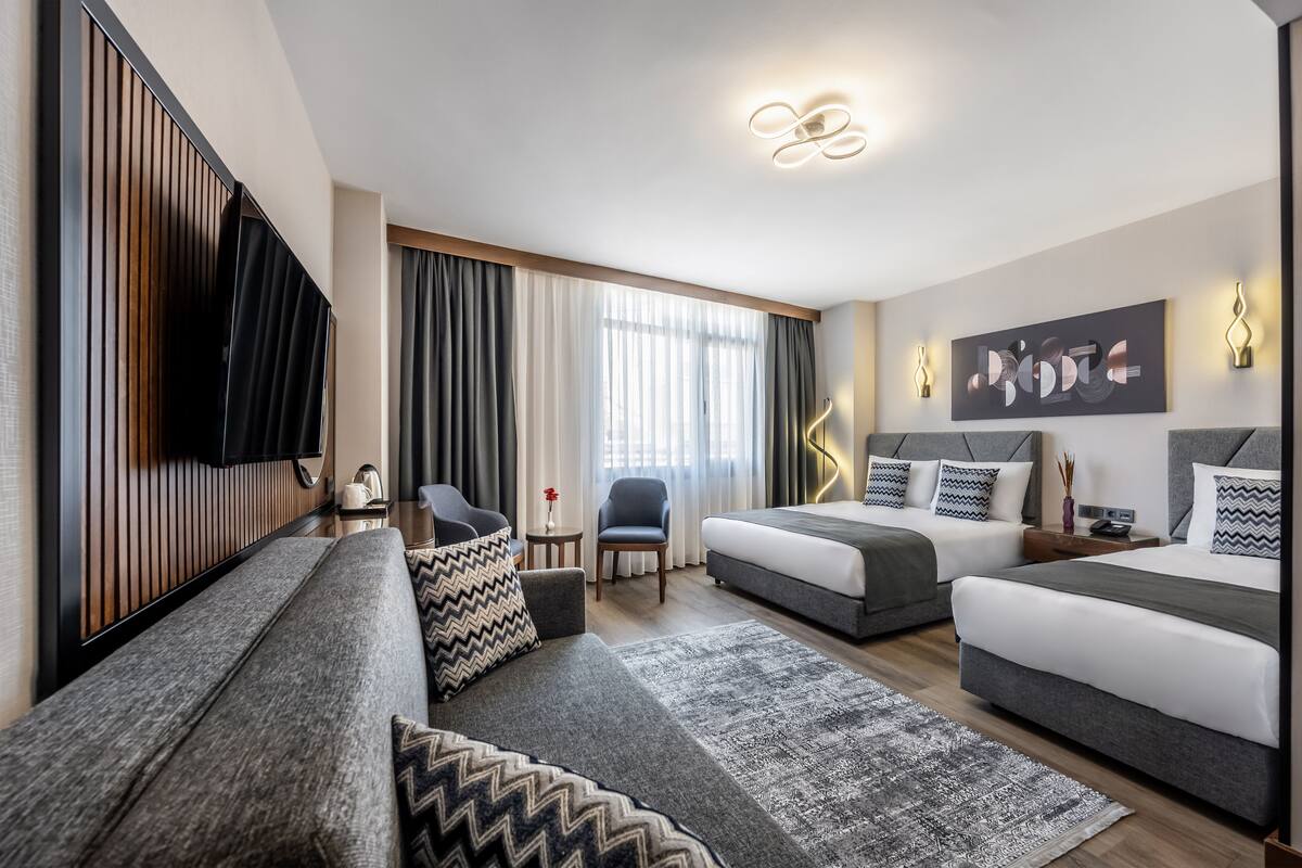 A modern hotel room with a sofa, two beds, a TV, a desk, and a large window with curtains.