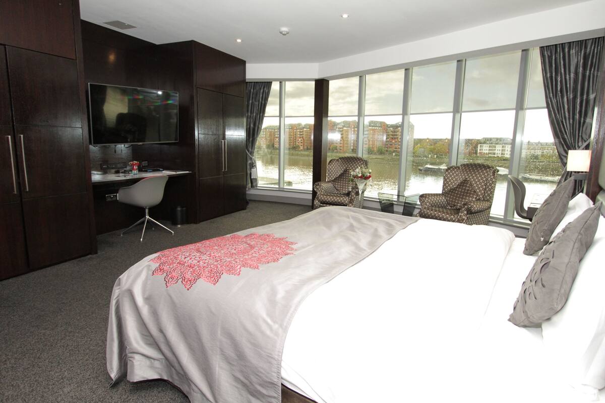 A modern hotel room with a large bed, a desk with a chair, a TV, and a view of the city through large windows.