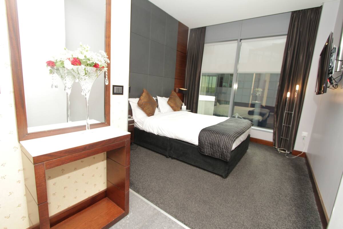 A hotel room with a large bed, a bedside table, a mirror, and a view of the city through large windows.