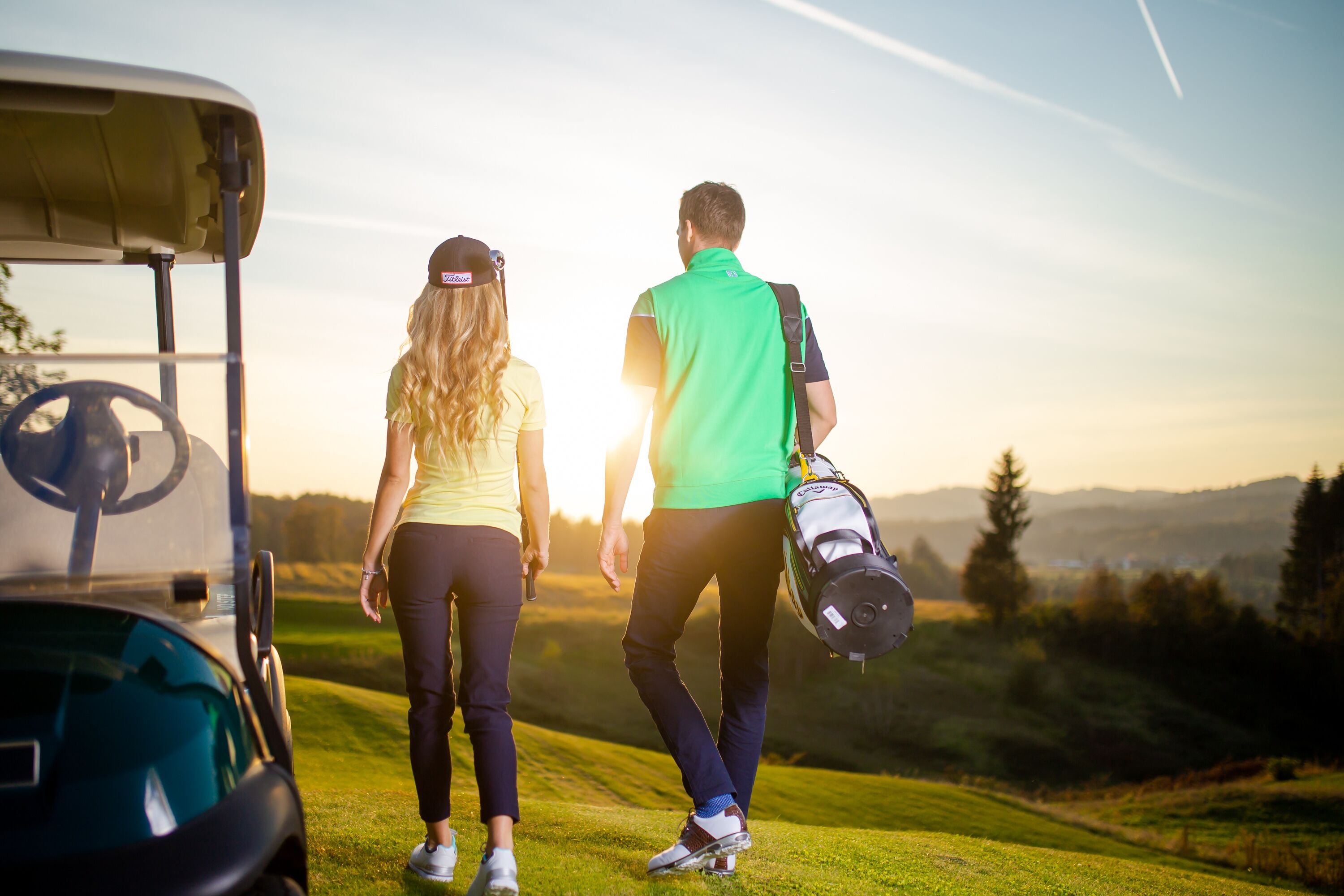Un carrito de golf y dos personas caminando en un campo de golf, con un atardecer de fondo.