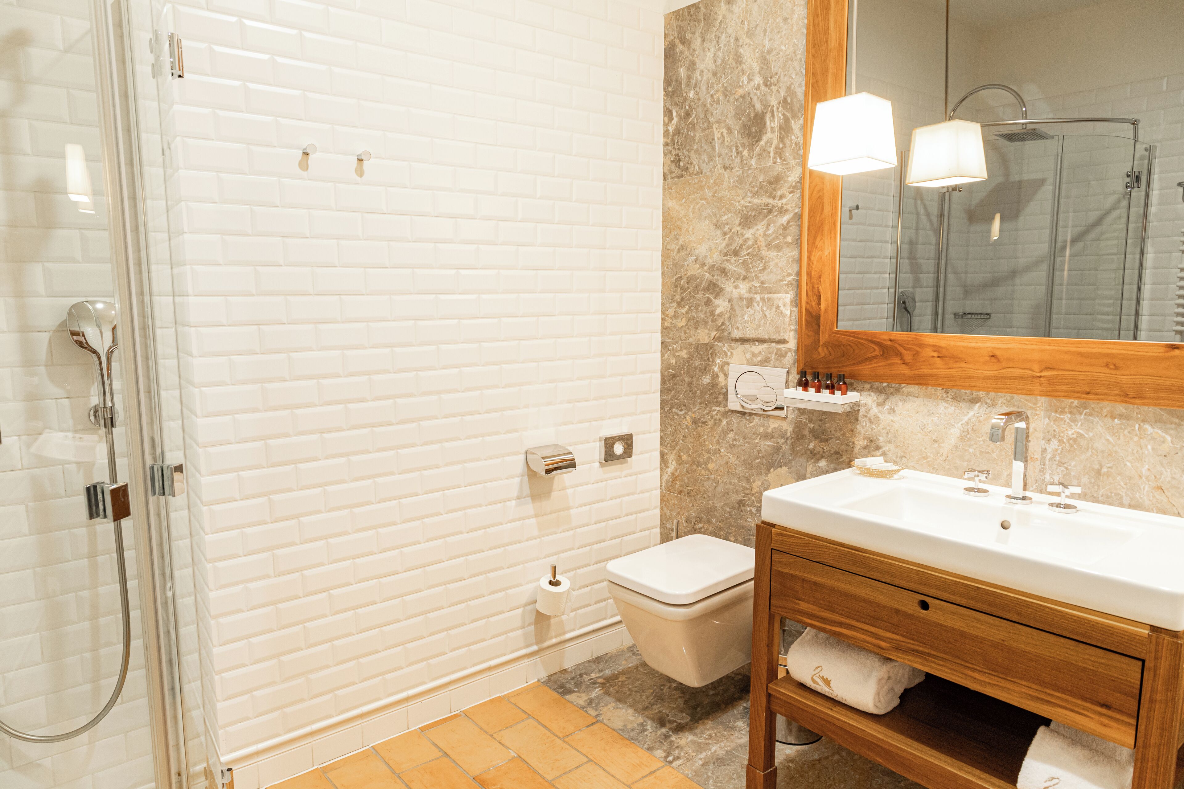 A bathroom with a glass shower enclosure, a white toilet, a wooden vanity with a mirror, and a marble backsplash.