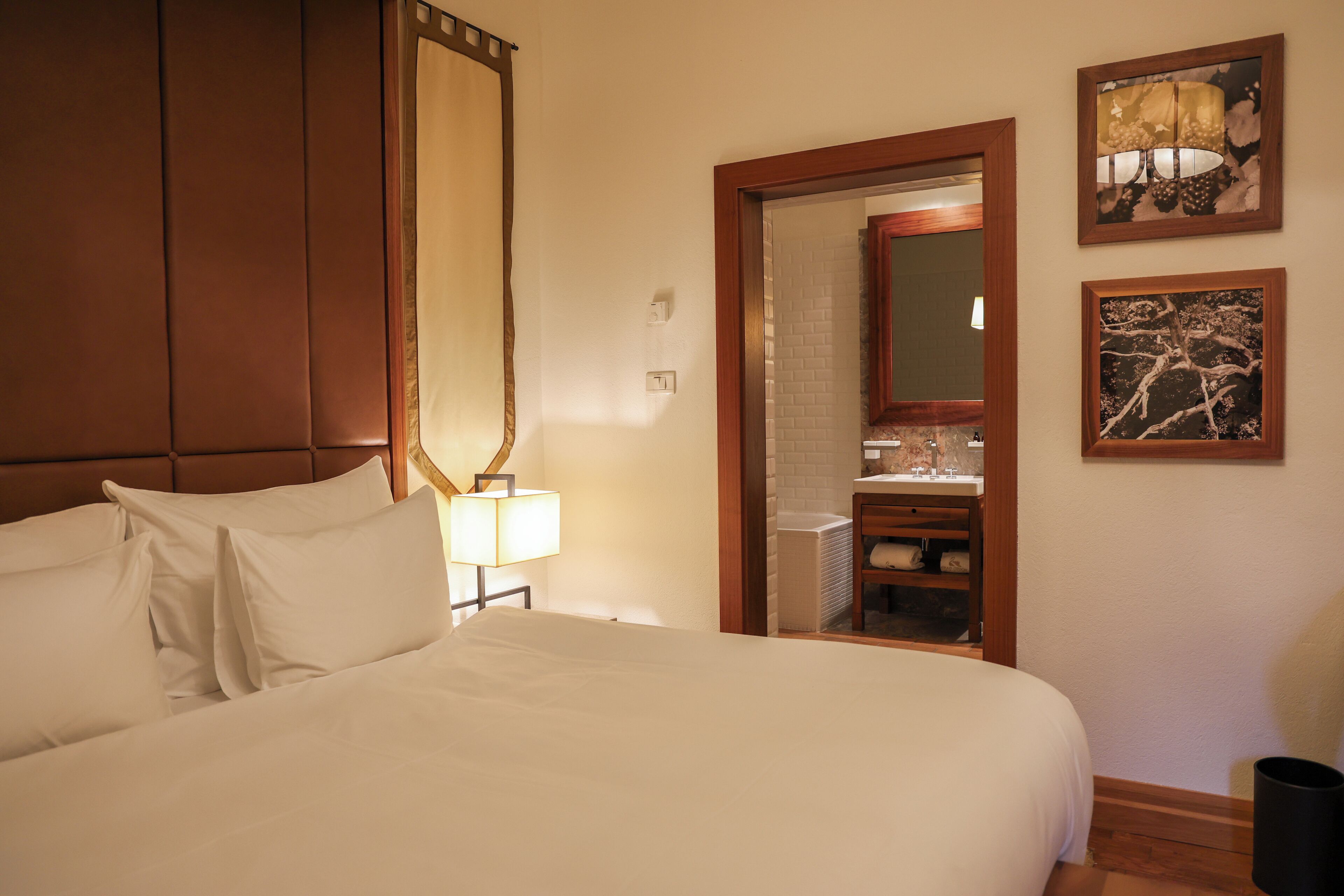 A hotel room with a large bed, a nightstand with a lamp, a wooden headboard, a bathroom visible through an open door, and two framed pictures on the wall.