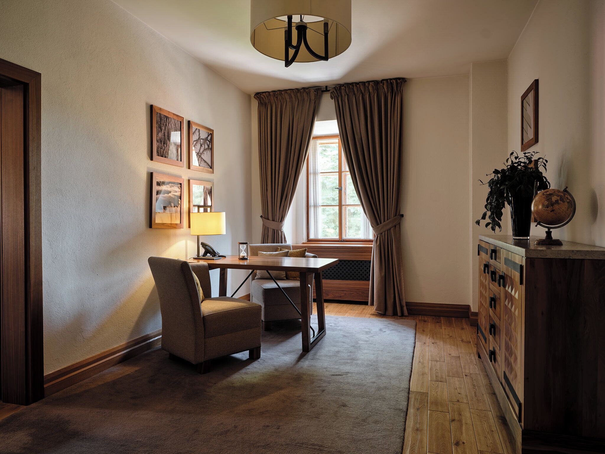 Ein gemütliches Zimmer mit Schreibtisch aus Holz, einem Sessel, einer Lampe, einem Globus und einer Pflanze auf einer Kommode.