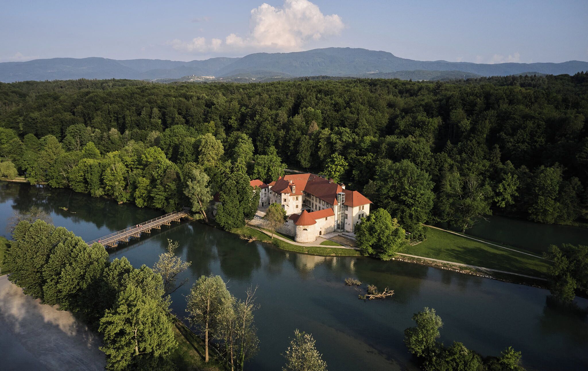 A castle-like building with a red roof, surrounded by dense forest and a lake.