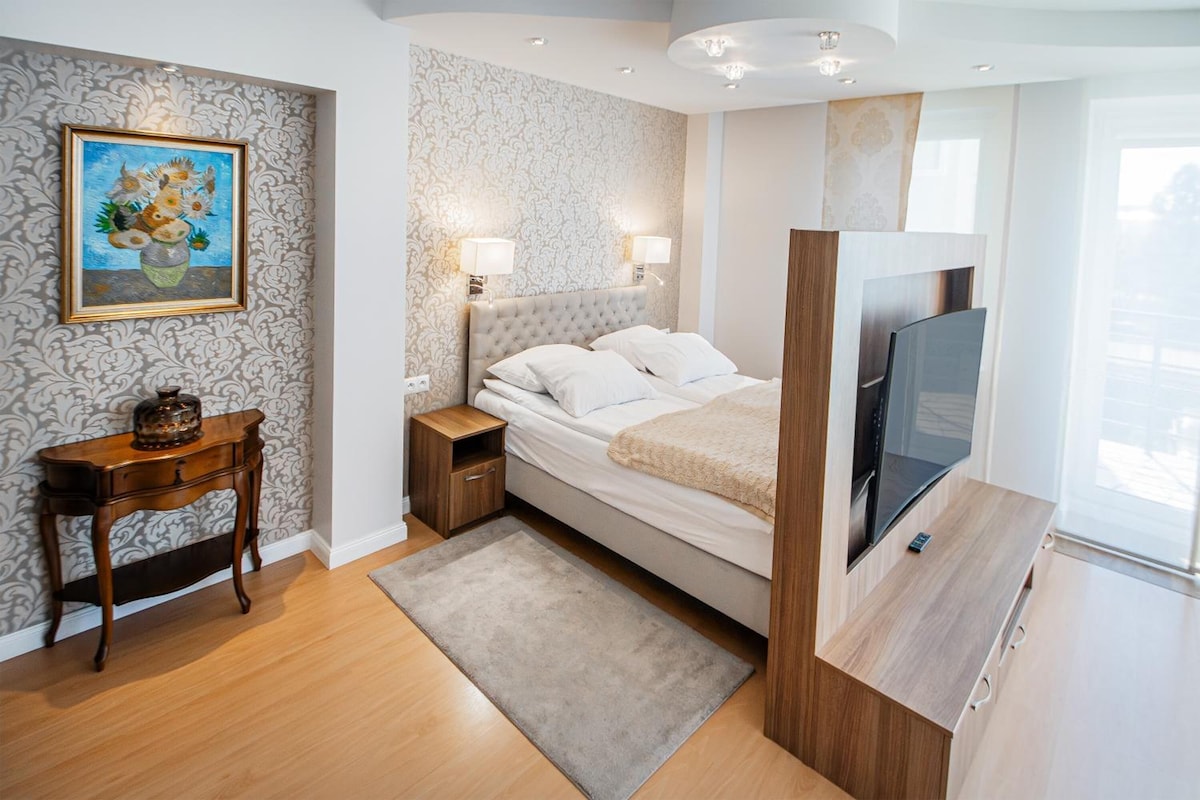 A modern bedroom with a bed, bedside lamps, a TV, a small table, and a painting on the wall.