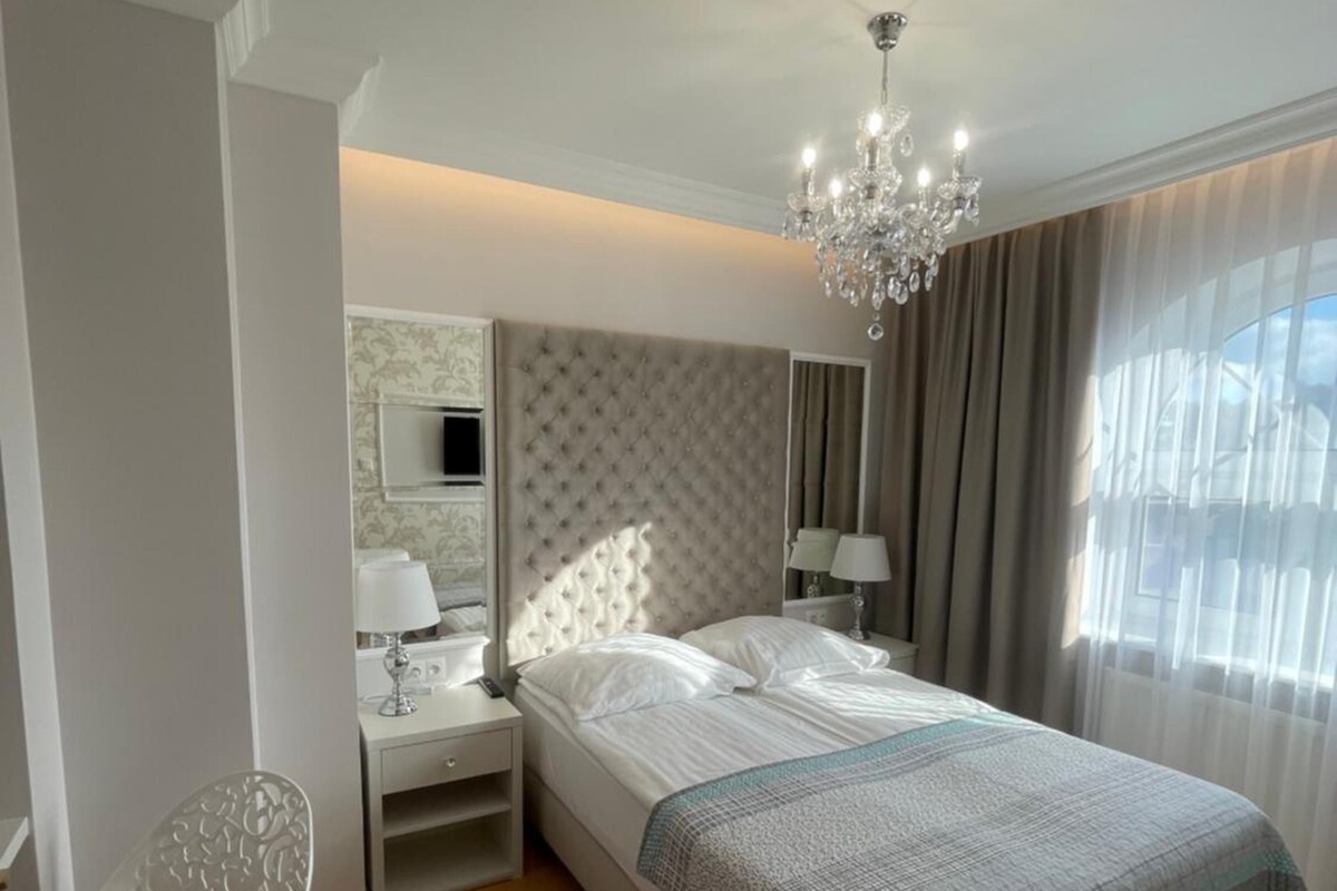 A bedroom with a bed, bedside tables, a chair, a chandelier, and a window with curtains.