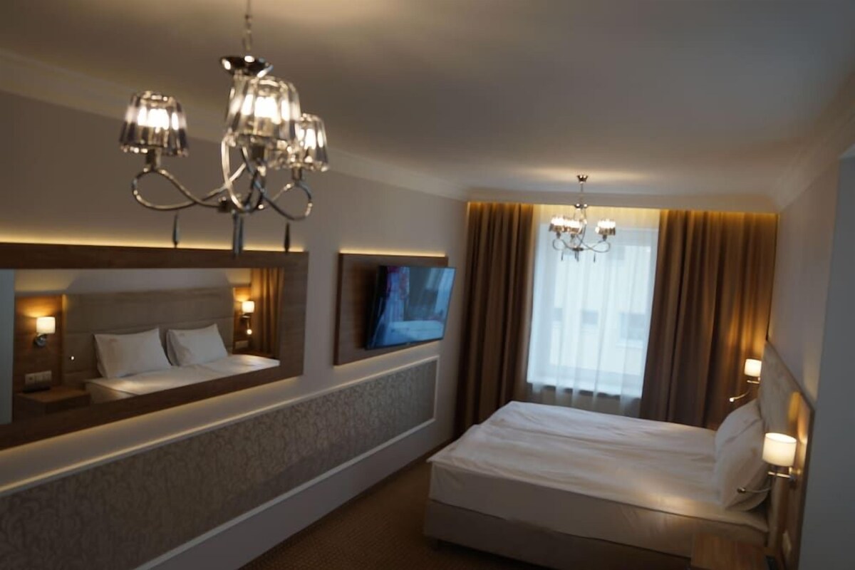 A hotel room with a large bed, a TV, a chandelier, and a window with curtains.