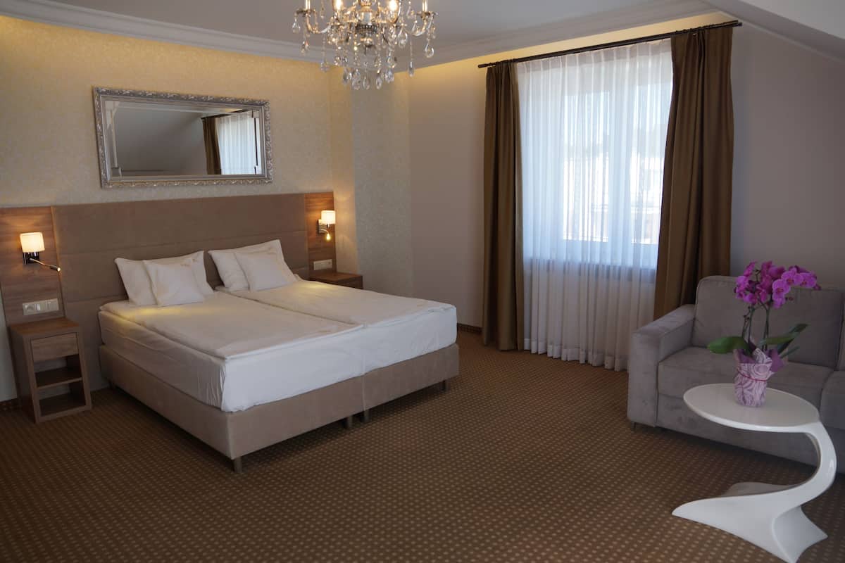 Quarto de hotel com uma cama grande, mesa de cabeceira, abajur, uma cadeira, pequena mesa com um vaso de flores e um lustre.