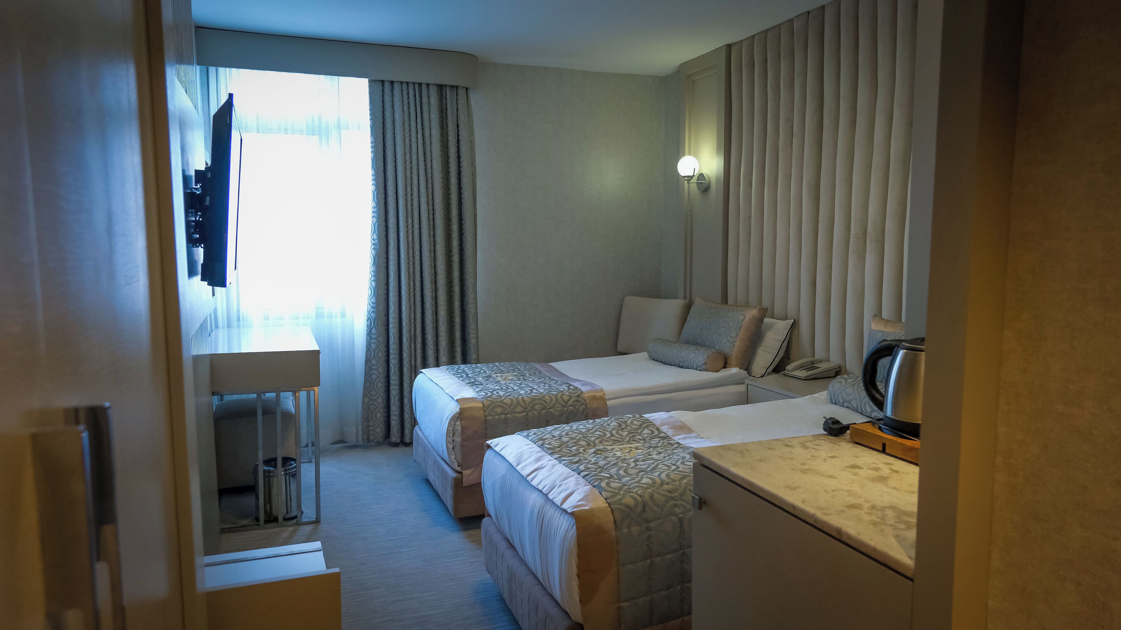 Habitación de hotel con dos camas, un escritorio, una silla, un cubo de basura, una lámpara y una tetera.