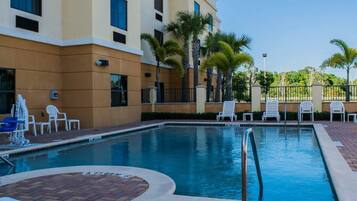 A swimming pool with lounge chairs and palm trees, surrounded by a building with multiple windows and a brick pathway.