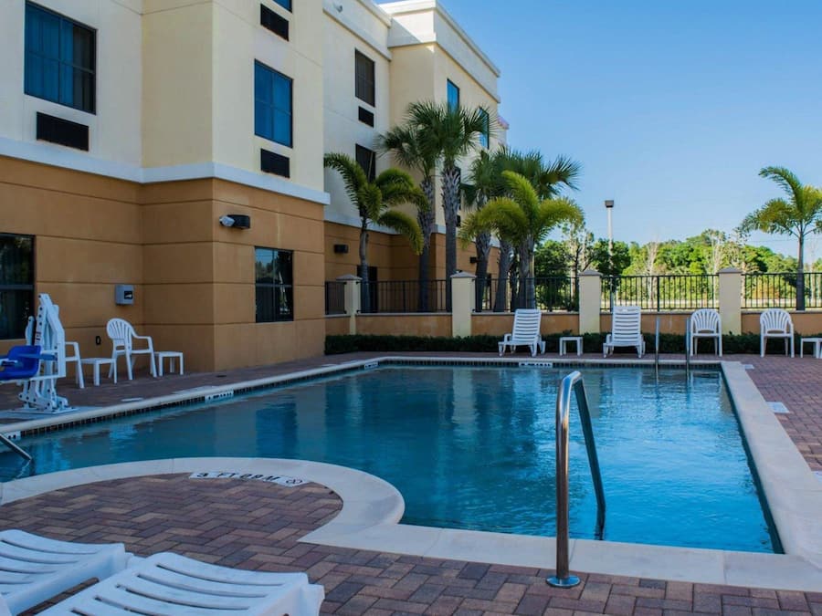 A swimming pool with lounge chairs and palm trees, surrounded by a building with multiple windows and a brick pathway.