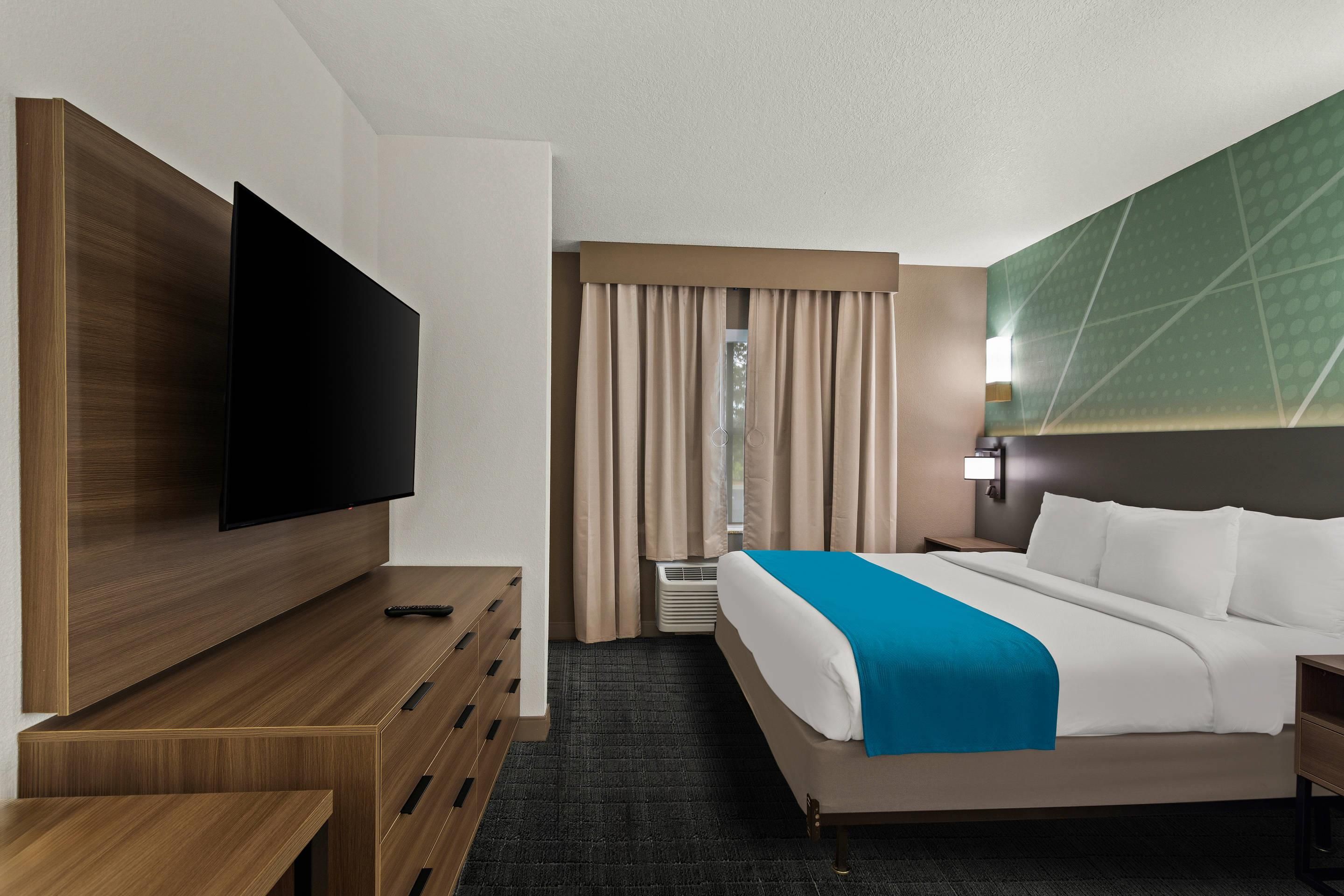 Een hotelkamer met een flatscreen televisie, een bed met wit en blauw beddengoed, een houten dressoir en een groot raam met gordijnen.