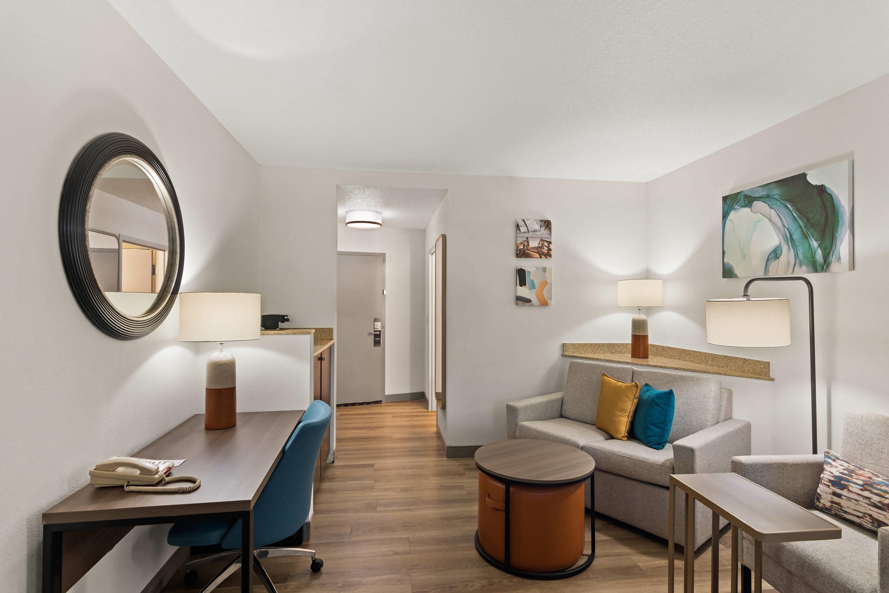 A hotel room with a desk, chair, sofa, and armchair. There are two lamps, a round table, and a small round table. The room has a mirror, artwork on the wall, and a door leading to another room.