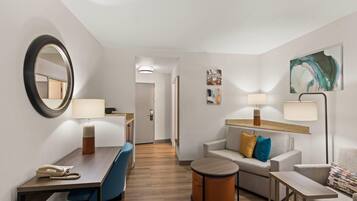 A hotel room with a desk, chair, sofa, and armchair. There are two lamps, a round table, and a small round table. The room has a mirror, artwork on the wall, and a door leading to another room.