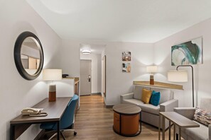 Een hotelkamer met een bureau, stoel, bank en fauteuil. Er zijn twee lampen, een ronde tafel en een klein rond tafeltje. De kamer heeft een spiegel, kunst aan de muur en een deur naar een andere kamer.