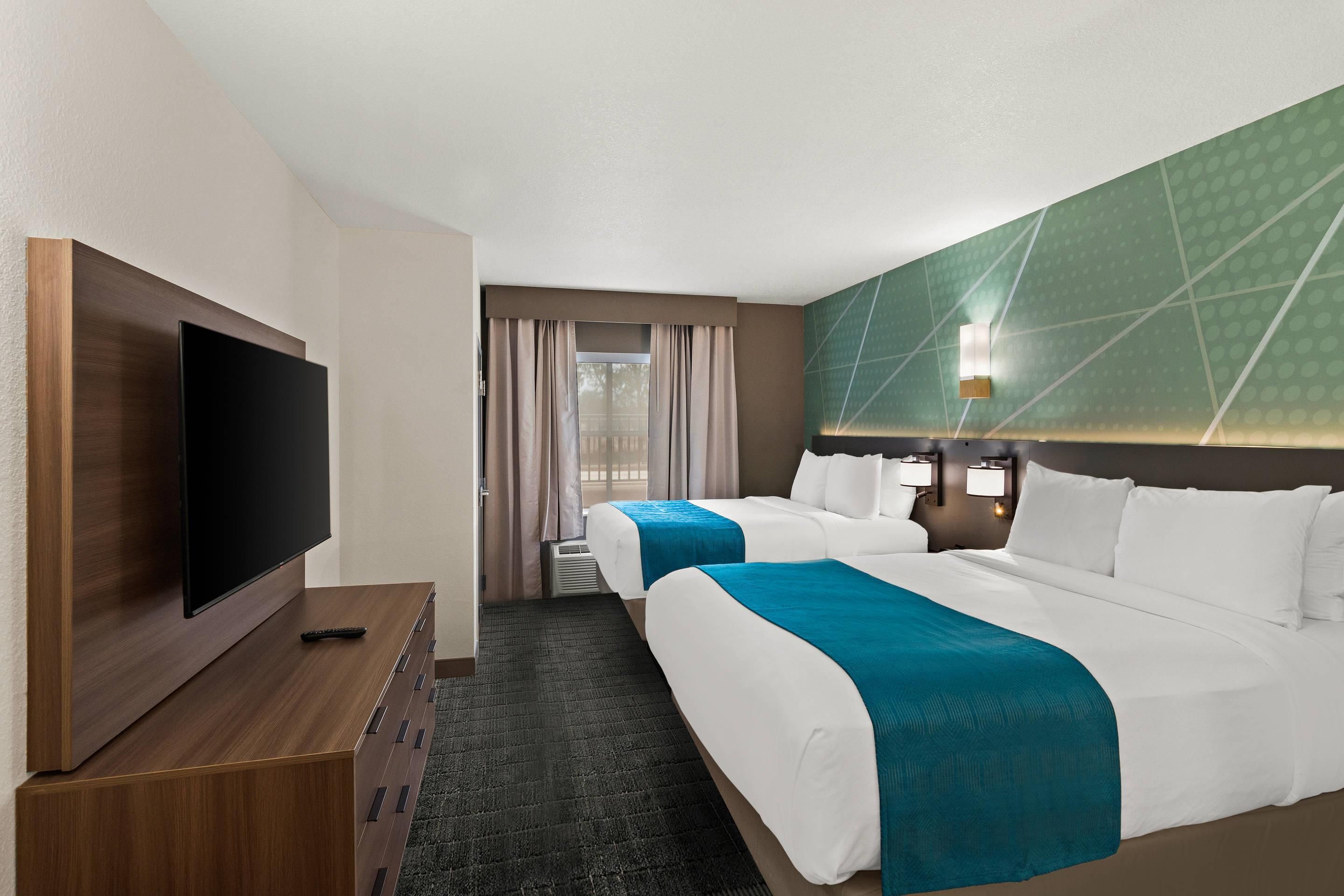 Habitación de hotel con televisor de pantalla plana, cómoda de madera, dos camas con ropa de cama blanca y verde azulada, pared con estampado verde y ventana con cortinas.