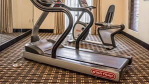Hotelkamer met een Star Trac loopband, een tweede fitnessapparaat, een aan de muur bevestigde televisie en een kamerbreed tapijt met een patroon.