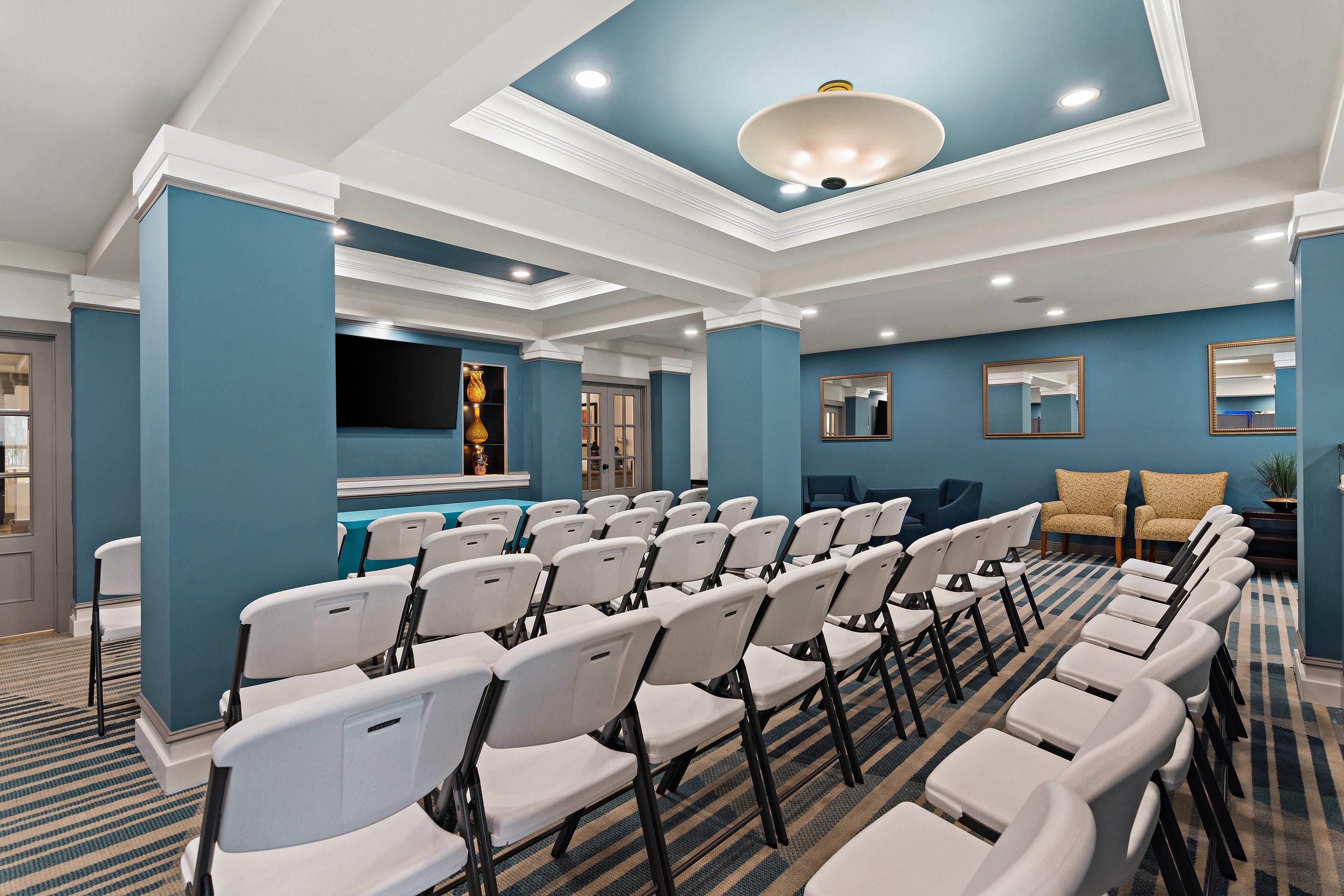 A conference room with rows of white chairs, a large flat-screen TV, and a ceiling with recessed lighting.