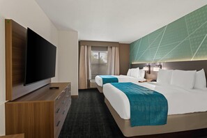 Een hotelkamer met een flatscreen-tv, een bureau met een afstandsbediening, twee bedden met blauwgroene dekbedden, een groot raam met gordijnen en een groen, geometrisch wandontwerp.