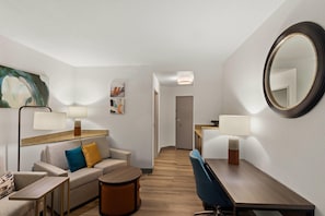 Habitación de hotel con sofá, una pequeña mesa redonda, una mesa de comedor, una silla, un espejo y dos lámparas fijadas a la pared.