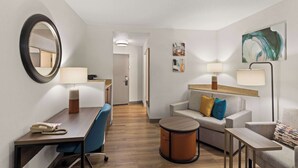 A hotel room with a desk, chair, sofa, and a small round table. There is a mirror on the wall, a lamp, and a painting.