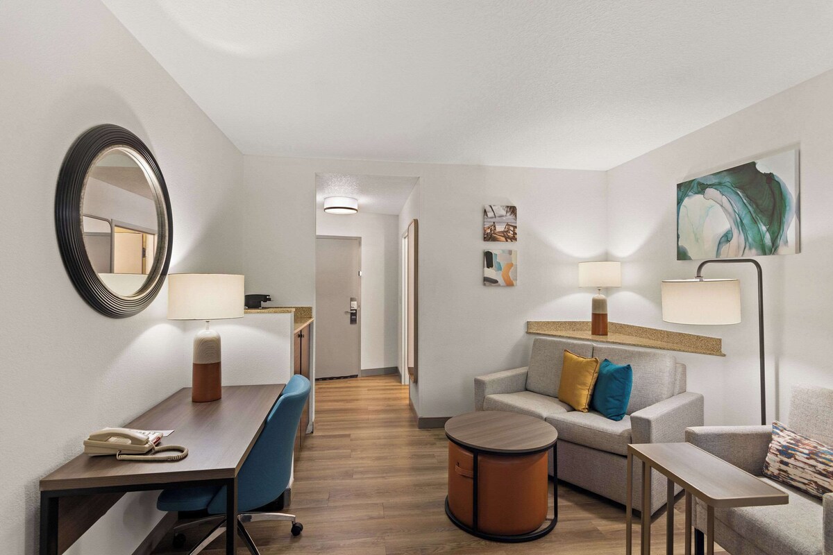 Ein Hotelzimmer mit Schreibtisch, Stuhl, Sofa und einem kleinen runden Tisch. An der Wand befindet sich ein Spiegel, eine Lampe und ein Gemälde.