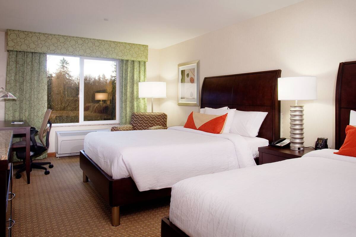 A hotel room with two beds, a desk, a chair, a TV, and a window with curtains.