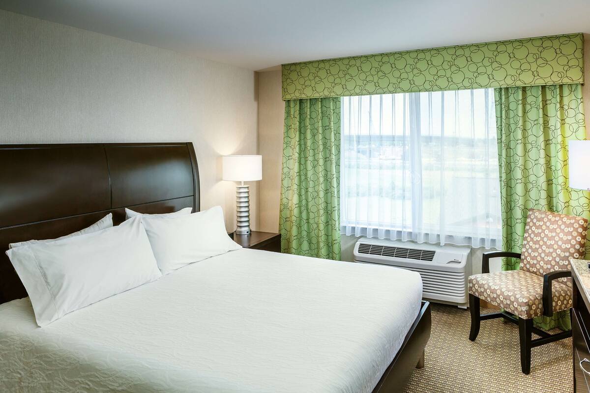 A hotel room with a large bed, a chair, a nightstand with a lamp, and a window with curtains.